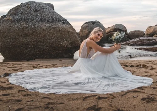 a bride in her wedding gown sitting on the beach and holding her bridal bouquet